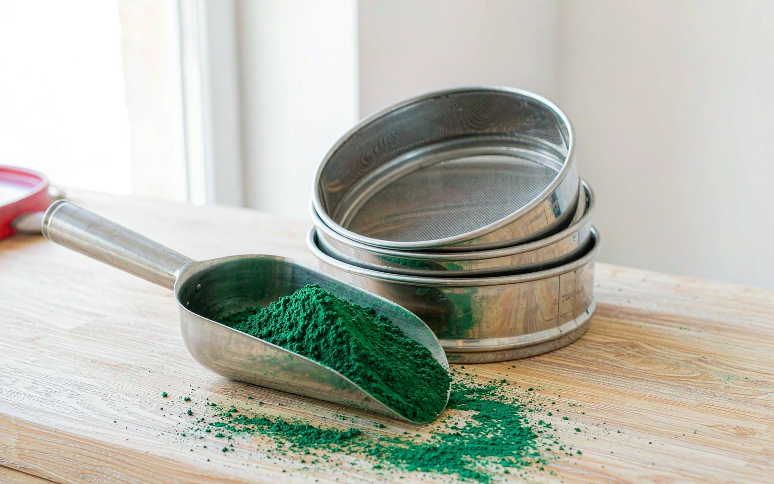 A stainless steel scoop of green vegetable powder beside a stack of test sieves on a workbench