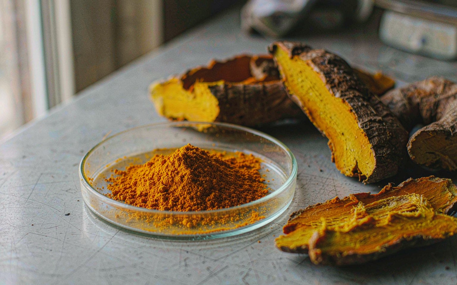 Bright orange yellow curcumin extract powder in a white dish beside split turmeric rhizome showing its deep orange interior