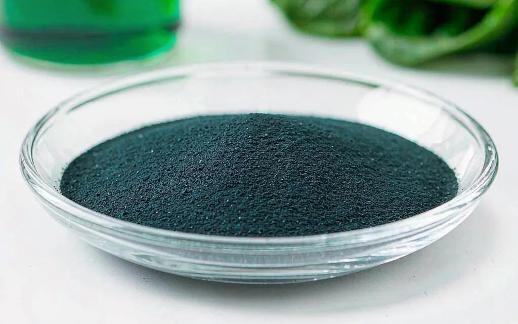 Sodium copper chlorophyllin: dark green to blue-black water-soluble powder in a glass dish, with a green solution and green leaves behind it
